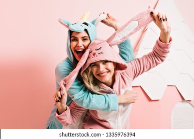 Ecstatic Girls In Kigurumi Fooling Around Together. Studio Shot Of Inspired Young Ladies Having Fun In Bedroom.