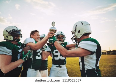 Ecstatic Group Of American Football Players Standing In A Huddle On A Field Raising A Championship Trophy In Celebration