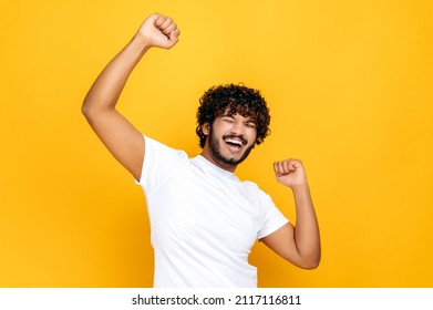 Ecstatic lucky happy curly Indian man, dancing, gesturing with fists, receives profit, glad to win lottery, standing on isolated orange background. Good news, success and happiness concept