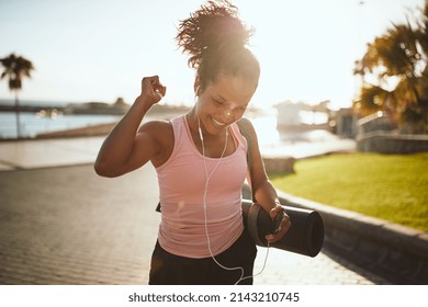 Ecstatic young African woman in sportswear listening to music on earphones and doing a fist pump while walking down the street carrying yoga matt - Powered by Shutterstock