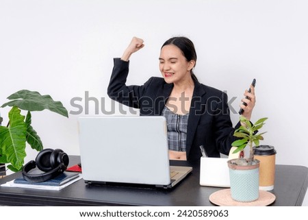 Ecstatic young woman at work raising her fist in triumph at her desk, surrounded by office essentials.