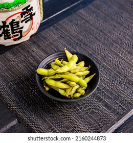 Edamame soybeans in the traditional Japanese ramen restaurant, with a black plate on a black mat and Japanese fonts sake barrel translation: Snow Crane