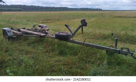 At The Edge Of A Barley Field In The Grass Stands A Car Carriage Trailer, Designed To Carry Plastic Or Aluminum Boats. It Has A Spare Tire And Planks Bolted To The Frame, Used For Off-roading