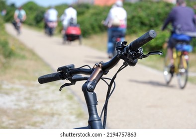 At the edge of a bike path: close-up of e-bike handlebars in the foreground, many cyclists, path and nature blurred in the background, landscape format, selective focus