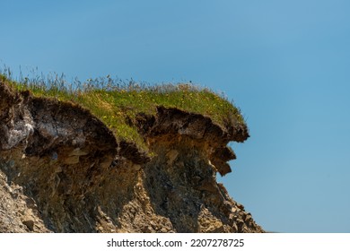 The Edge Of A Grassy Cliff With The Sod Hanging Over The Soil. The Ground Of The Hillside Is In The Shape Of A Wave. The Rugged Coastline Land Has Eroded Under The Sod. The Background Is A Blue Sky.