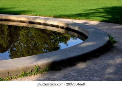 edge of a stone sandstone circular fountain in the park. built of sandstone filled with water. lined with a light threshing gravel road with a border of granite cubes