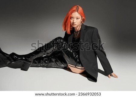 edgy style, asian woman with red hair sitting in bold outfit and latex boots on grey background