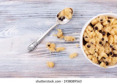 Edible Cookie Dough In A Cup With The Spoon And Crumbs On The White Wooden Background, Close Up.