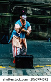 Edinburgh, United Kingdom, 3 Of August 2018 A Street Artist Talking And Raising A Hand Dressed As An Asian Person And With Makeup Doing A Show With A Black Suitcase In The City Center