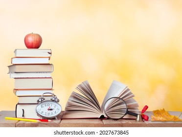 education background with stack of books, red apple, colored pencils, alarm clock, magnifying glass, golden bell, opened book and maple leaf on the wooden table with copy space