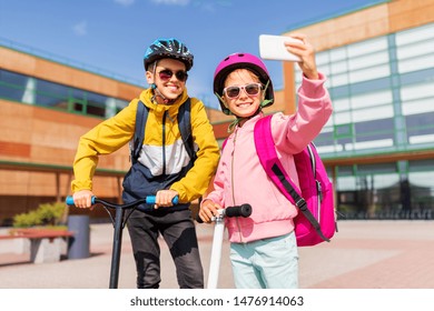 education, childhood and people concept - happy school children in helmets with backpacks riding scooters and taking selfie by smartphone outdoors