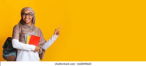 Education Concept. Happy black muslim student girl in hijab with notepads and backpack pointing aside at copy space over yellow background, panorama