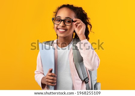 Education Concept. Portrait of positive smiling African American girl touching glasses holding textbooks, wearing backpack and looking at camera isolated on yellow studio background