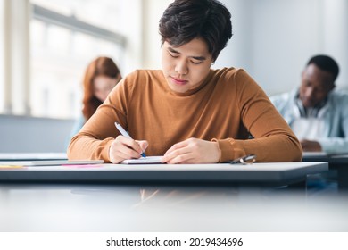 Education Concept. Portrait of serious asian male student sitting at desk in classroom at university, writing in notebook, taking notes, exam or test. Return to college and high school