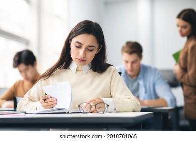 Education Concept. Portrait Of Serious Female Student Sitting At Desk In Classroom At University, Writing In Notebook, Taking Notes, Exam Or Test. Return To College And High School