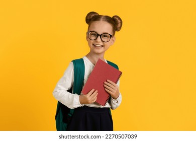 Education Concept. Portrait Of Smiling Cute Schoolgirl With Backpack And Books In Hands, Cheerful Little Female Pupil Wearing Eyeglasses Standing Isolated Over Yellow Studio Background, Copy Space