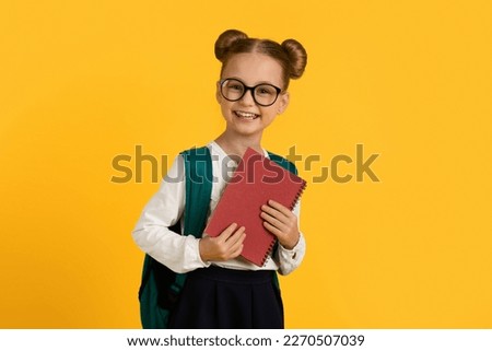 Education Concept. Portrait Of Smiling Cute Schoolgirl With Backpack And Books In Hands, Cheerful Little Female Pupil Wearing Eyeglasses Standing Isolated Over Yellow Studio Background, Copy Space