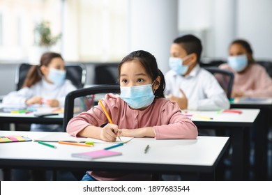 Education During Pandemic. Asian Girl Sitting At Table In Classroom At School Or Kindergarten, Wearing Surgical Face Mask, Writing Or Drawing In Notebook. New Normal, Safe Learning