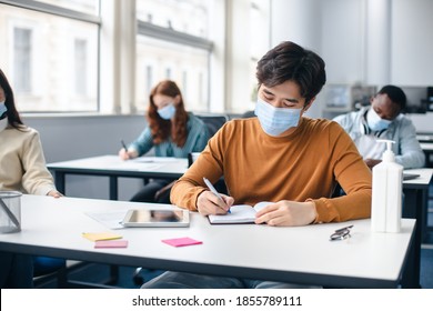 Education During Pandemic. Asian Male Student Sitting At Table In Classroom At High School, Wearing Surgical Face Mask, Writing Test Or Taking Notes In Notebook, Sanitizer Bottle On The Desk