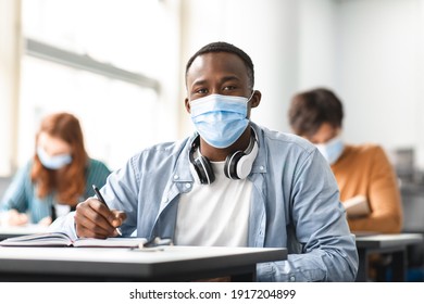 Education During Pandemic Concept. Portrait Of Black Male Student Sitting At Table In Classroom At University, Wearing Protective Medical Face Mask, Writing In Notebook, Looking Posing At Camera