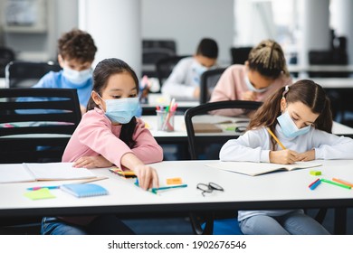 Education During Pandemic. Diverse Girls Sitting At Table In Classroom At School Or Kindergarten, Wearing Disposable Surgical Face Mask, Writing Or Drawing In Notebook. New Normal, Safe Learning