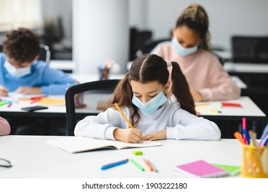 Education During Pandemic. Small Girl Sitting At Desk In Classroom At School Or Kindergarten, Wearing Disposable Surgical Face Mask, Writing Or Drawing In Notebook. New Normal, Safe Learning