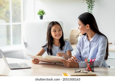 Education and early development concept. Korean mother helping her daughter with homework, sitting in kitchen interior, copy space. Happy asian girl holding book and smiling to mom