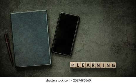 Education E-learning Concept, Blank Screen Smart Phone With Notbook And Word E-learning On Wood Cube Block On Black Table Background, Flat Lay Top View Copy Space.