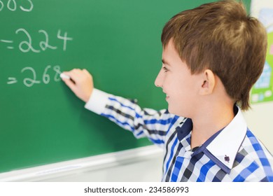 education, elementary school, learning, math and people concept - little smiling schoolboy writing numbers on green chalk board in classroom