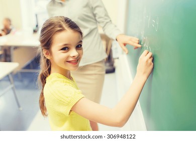 Education, Elementary School, Learning, Math And People Concept - Little Smiling Schoolgirl Writing Numbers On Green Chalk Board In Classroom