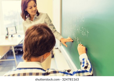 education, elementary school, learning, math and people concept - close up of little schoolboy with teacher writing on chalk board and solving task in classroom