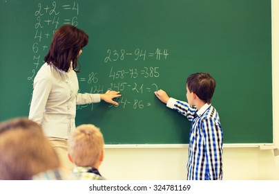 education, elementary school, learning, math and people concept - close up of little schoolboy with teacher writing on chalk board and solving task in classroom