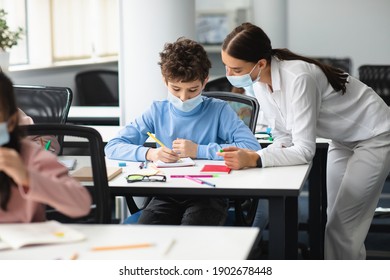 Education, Elementary School, Learning And People Concept. Female Teacher In Disposable Protective Mask Helping Small School Boy Who Sitting At Desk In Classroom, Standing Near Table, Explaining Task