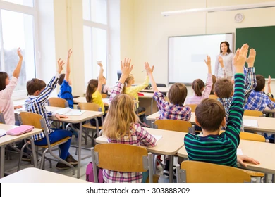 Education, Elementary School, Learning And People Concept - Group Of School Kids With Teacher Sitting In Classroom And Raising Hands