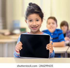 Education, Elementary School, Technology, Advertisement And Children Concept - Little Student Girl Showing Blank Black Tablet Pc Computer Screen Over Classroom And Classmates Background