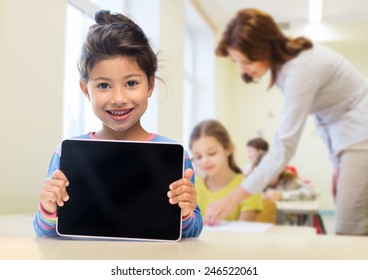 Education, Elementary School, Technology, Advertisement And Children Concept - Little Student Girl Showing Blank Black Tablet Pc Computer Screen Over Classroom And Classmates Background