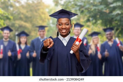 Education, Graduation And People Concept - Happy Graduate Student Woman In Mortarboard And Bachelor Gown With Diploma Pointing Finger To Camera Over Group Of Bachelors At Park On Background