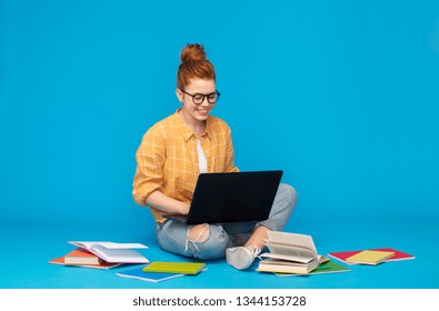 education, high school, technology and people concept - red haired teenage student girl in checkered shirt and torn jeans with books using laptop computer over bright blue background