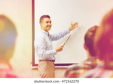Education, High School, Technology And People Concept - Smiling Teacher With Notepad, Laptop Computer Standing In Front Of Students And Showing Something On White Board In Classroom