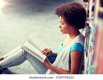 education, high school, university, learning and people concept - african american student girl reading book sitting on floor at library