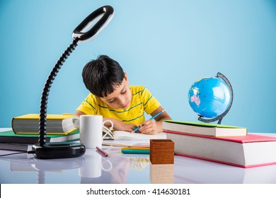 Education at home concept - Cute little Indian/Asian boy studying or completing home work on study table with pile of books, educational globe, laptop computer, coffee mug etc