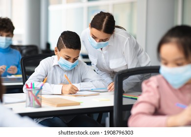 Education, Junior School, Guidance Concept. Female teacher in protective face mask helping small schoolboy who is sitting at table in classroom, standing near table, pupil writing test