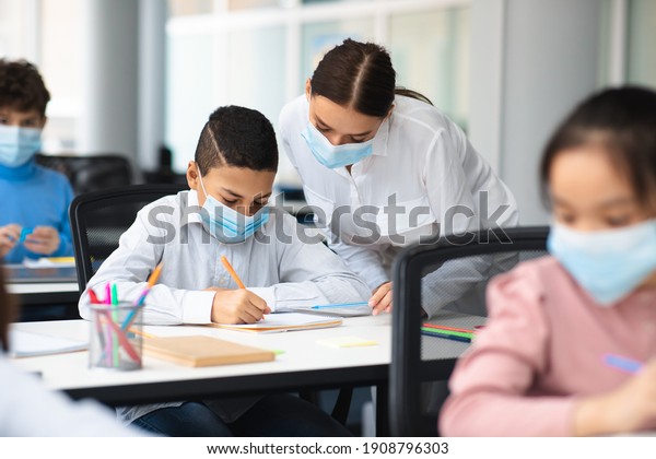 Education, Junior
School, Guidance Concept. Female teacher in protective face mask
helping small schoolboy who is sitting at table in classroom,
standing near table, pupil writing
test