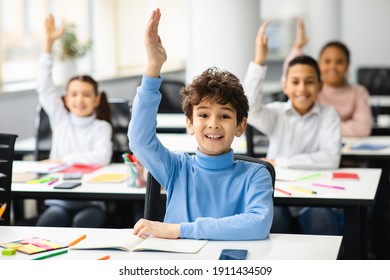 Education, Junior School, Learning and People Concept. Happy schoolboy raising hand for an answer, sitting at desk in classroom with diverse group of smiling classmates, studying with pleasure - Powered by Shutterstock