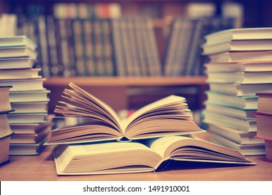 Education Learning Concept With Opening Book Or Textbook In Old Library, Stack Piles Of Literature Text Academic Archive On Reading Desk And Aisle Of Bookshelves In School Study Class Room Background