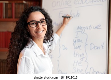 Education And Learning Concept. Portrait of smiling female teacher standing at whiteboard, explaining grammar rules to students. Excited woman in glasses looking at camera, writing on the board - Shutterstock ID 1841911015