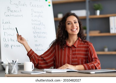 Education And Learning Concept. Portrait of smiling female teacher pointing at whiteboard, explaining English grammar rules to students. Excited woman looking at camera, sitting at desk - Powered by Shutterstock