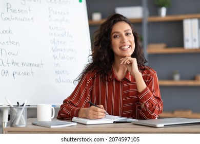 Education And Learning Concept. Portrait Of Smiling Beautiful Curly Female English Teacher Sitting At Desk Near Whiteboard With. Happy Lady Posing And Looking At Camera, Writing In Notebook