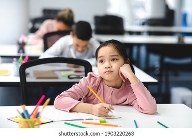 Education And Learning Concept. Portrait of tired and bored small asian girl sitting at desk in classroom at school, writing in notebook and thinking, looking away at window, resting head on hand - Powered by Shutterstock