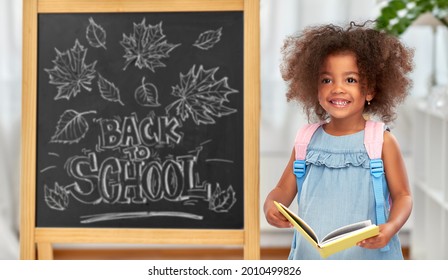 Education, Learning And People Concept - Happy Little African American Girl With Book And Backpack Over Chalkboard With Back To School Lettering On Background